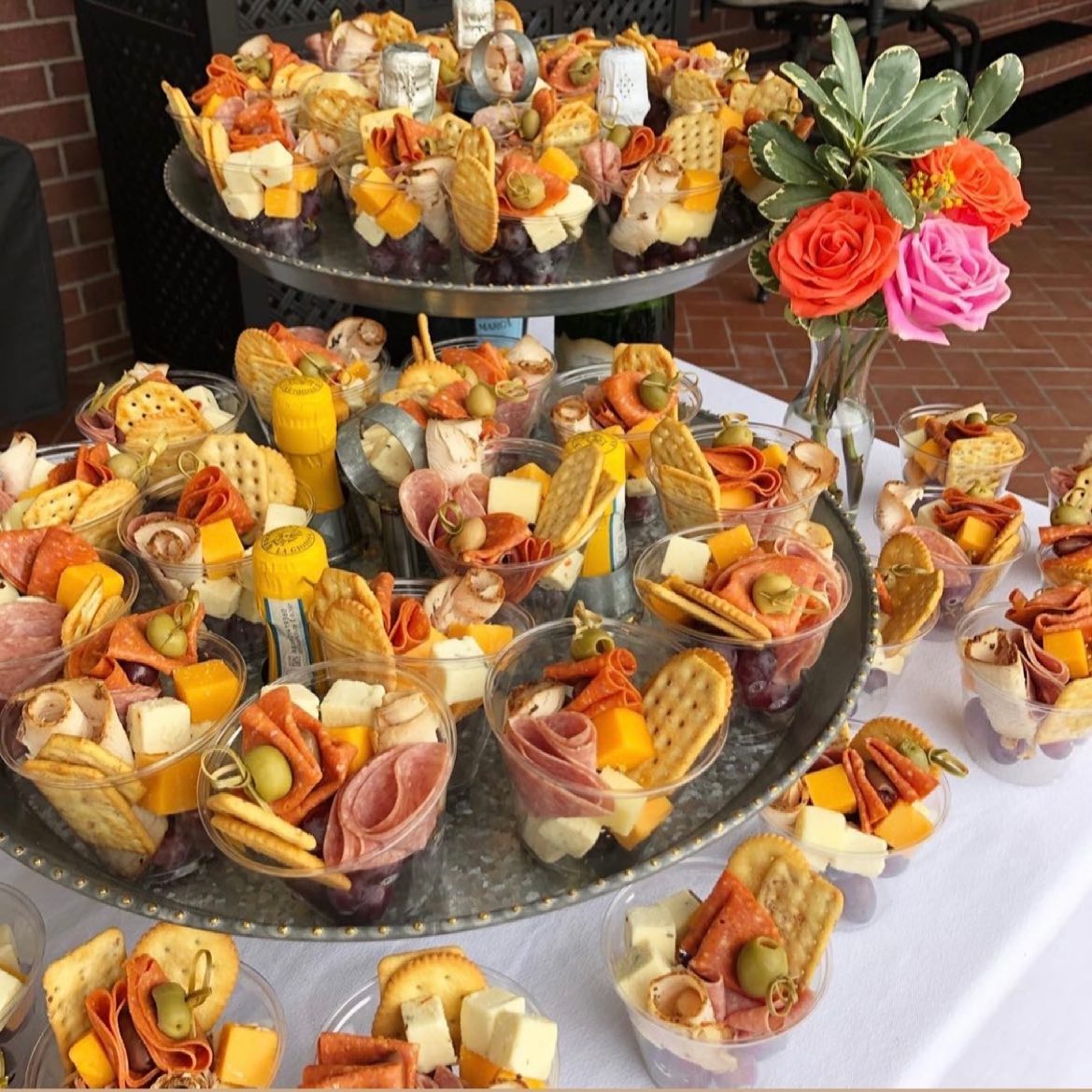 cups full of meat, fruit, vegetables, cheese and crackers on platters with flowers in the background